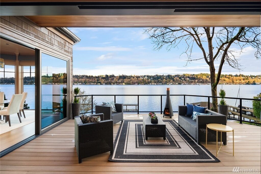 13.1725440_11_1 Modern outdoor patio with black wicker furniture and a patterned rug overlooks a scenic lake. Designed by a residential architect, a cozy fireplace and potted plants enhance the space, while large windows connect it to the dining area inside.