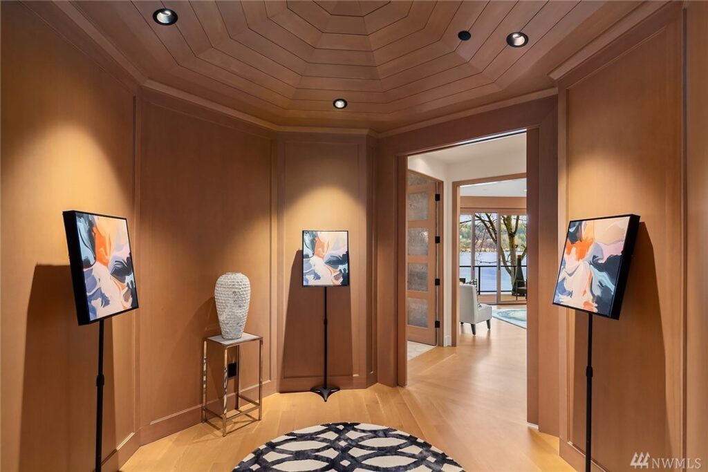 15.11725440_20_0 A modern hallway by a residential architect features wood-paneled walls, a patterned rug, abstract art on stands, and a sculpted vase on a small table; open double doors reveal a view of the lake and trees outside.