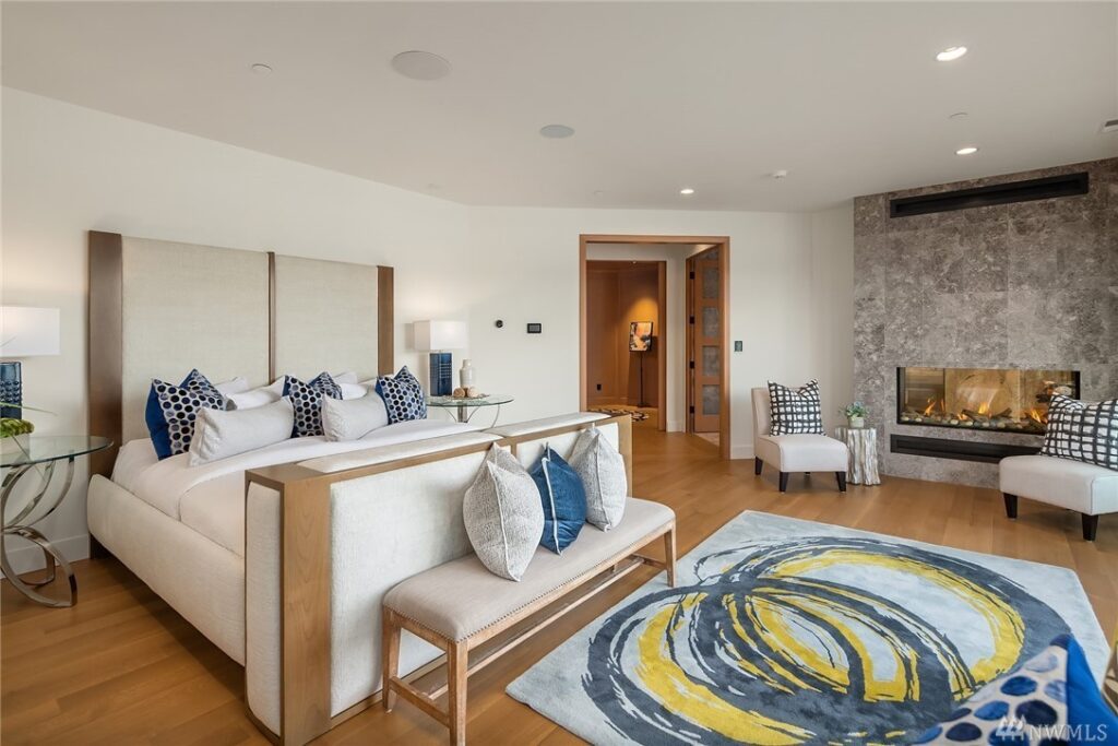 15.21725440_27_1 Spacious modern bedroom designed by a residential architect, featuring wood flooring, a large bed with white and blue pillows, an abstract blue and yellow rug, chairs by a stone fireplace, and neutral walls with minimal decor.