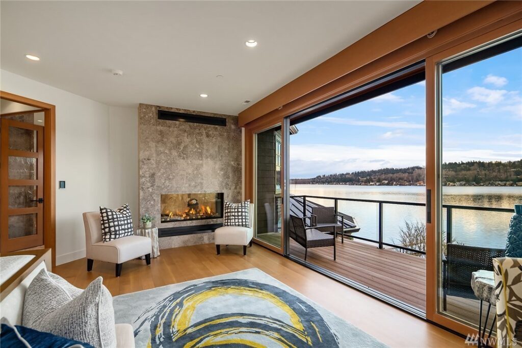 16.1725440_26_1 Modern living room designed by a residential architect, featuring a fireplace, two chairs, and large sliding glass doors opening to a balcony with a scenic lakeside view beneath a partly cloudy sky. Wooden floors and contemporary decor complete the space.