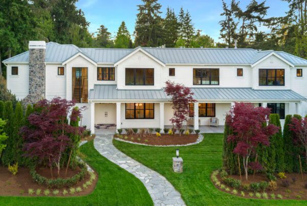 A large, modern two-story white house designed by a residential architect, featuring a metal roof, stone chimney, and many windows. Surrounded by lush lawn, landscaped gardens, and tall trees with a stone path leading to the entrance.