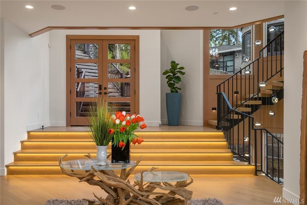 2.1725440_12_1 Bright modern entryway designed by a residential architect, featuring wood double doors, a glass staircase with black railing, illuminated steps, a tall potted plant, and a round glass table with driftwood base and red tulips.