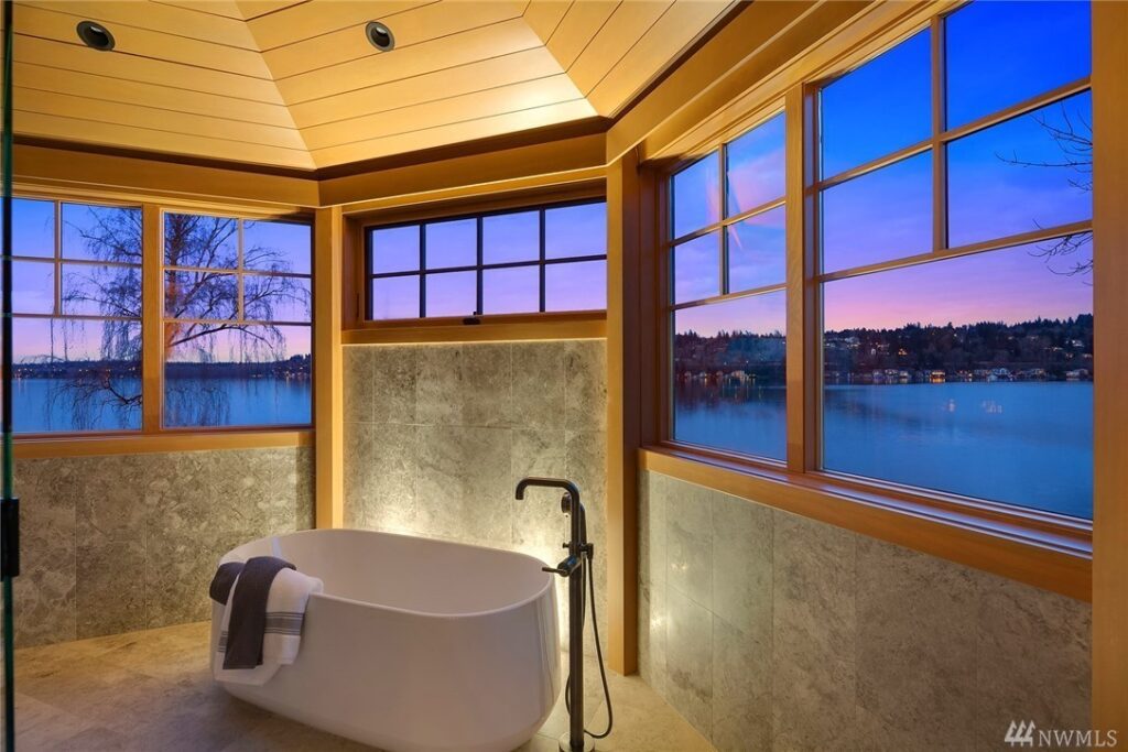 21.1725440_21_1 A modern bathroom, designed by a residential architect, features a freestanding tub with floor-mounted faucet and large windows overlooking a serene lake and tree-lined shore at sunset. Towels are neatly placed on the tub’s edge.