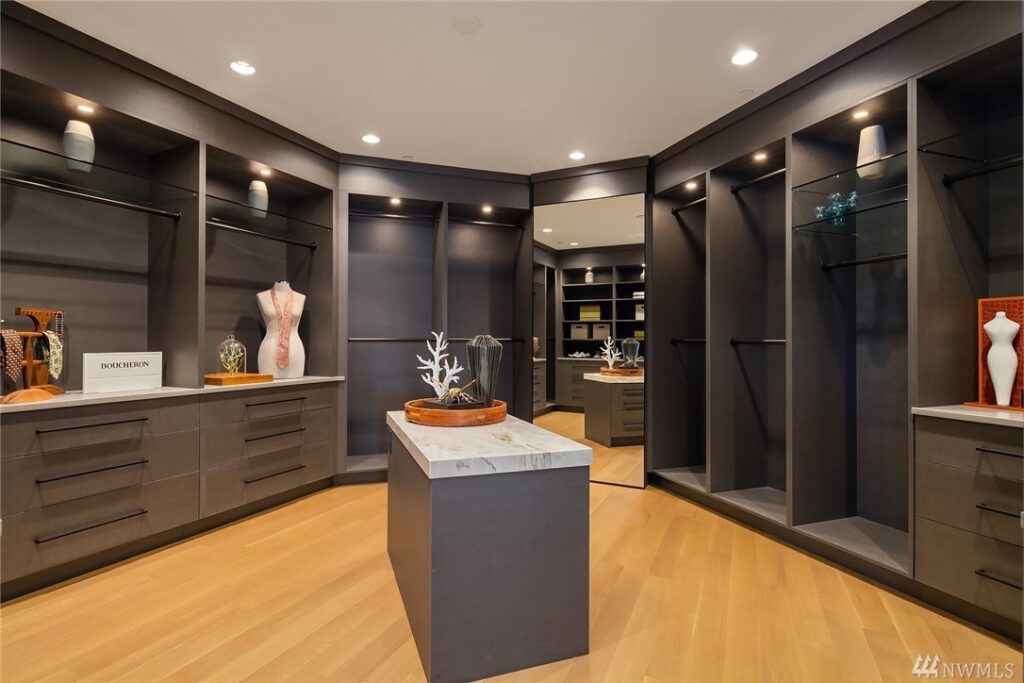 22.1725440_28_1 Spacious walk-in closet designed by a residential architect, featuring dark built-in shelves, drawers, display mannequins, decorative items, wood flooring, and a central island with a marble top—all under recessed lighting.