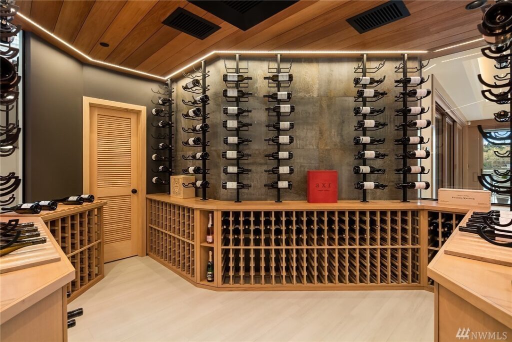 27.1725440_31_0 Modern wine cellar designed by a residential architect, featuring wooden racks and shelves, bottles stored horizontally on wall-mounted metal holders, a red wine box in the center, and warm lighting that enhances the inviting space.