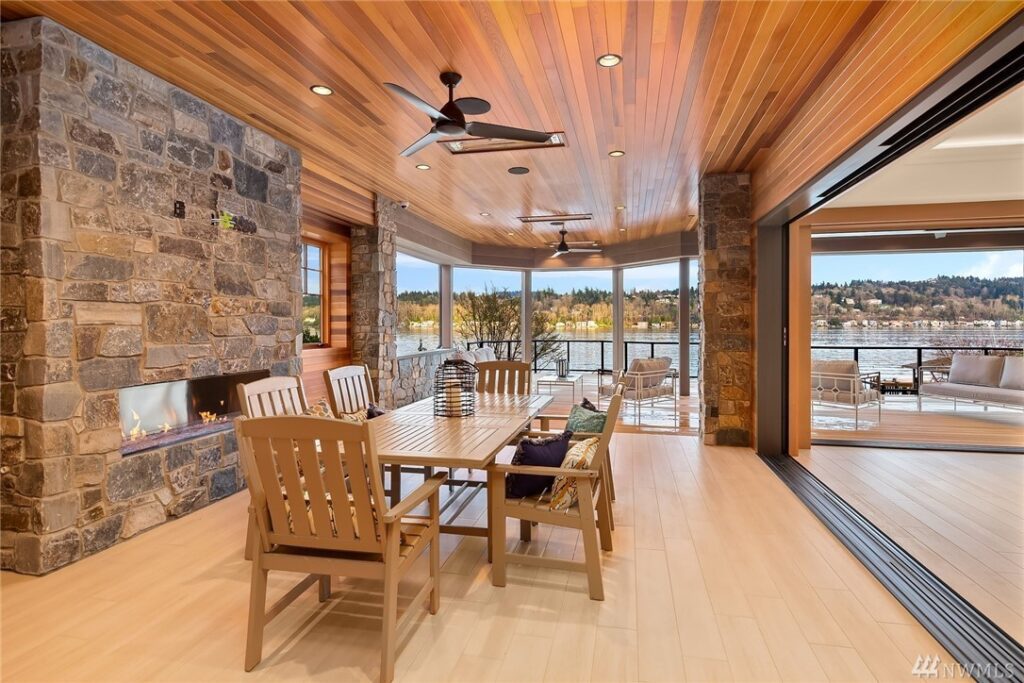 32.1725440_35_1 A spacious covered patio, designed by a residential architect, features a wood ceiling, stone fireplace, ceiling fans, and dining table—overlooking a scenic lake and trees through large open windows.