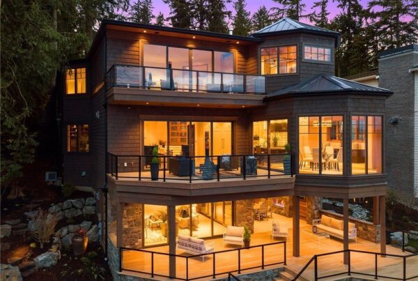A modern, multi-story house designed by a skilled residential architect features large glass windows and balconies, warmly illuminated at dusk and surrounded by tall trees and outdoor stone landscaping.