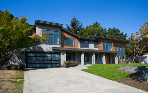 Modern two-story house designed by a residential architect, featuring large windows, a double garage with glass doors, and a spacious driveway. Green lawn and trees surround the home under a clear blue sky.