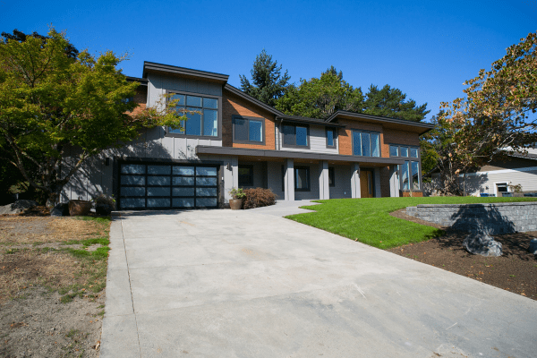 Modern two-story house designed by a residential architect, featuring large windows, a double garage with glass doors, and a spacious driveway. Green lawn and trees surround the home under a clear blue sky.