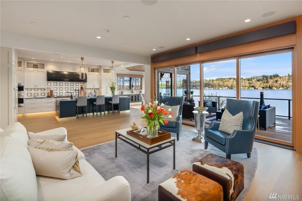 4.1725440_2_0 Modern open-concept living room designed by a residential architect features a white sofa, blue armchairs, coffee table with flowers, and large windows with lake views. The space flows into a kitchen with bar seating and outdoor patio access.