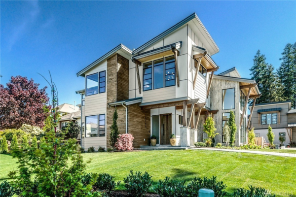 Modern two-story house, designed by a skilled residential architect, features large windows, a mix of wood and light-colored siding, landscaped lawn with shrubs and trees, all set under a clear blue sky.