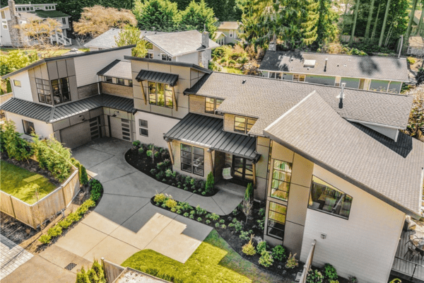 Aerial view of a modern two-story house, designed by a residential architect, with large windows, dark roof, and landscaped yard. A driveway curves toward the garage, surrounded by trees and neighboring homes.
