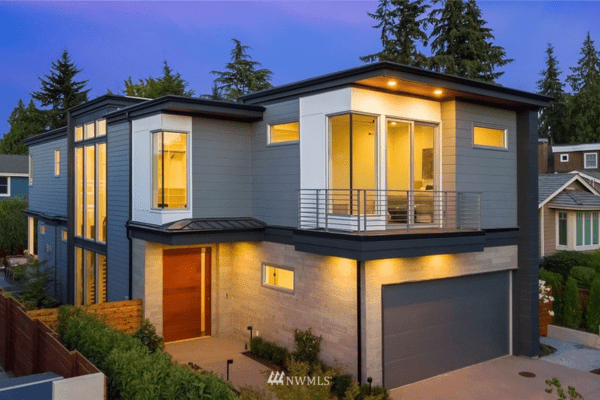 Modern two-story house with gray siding, large windows, a balcony, and exterior lights illuminating the driveway and entrance at dusk. Designed by a residential architect, it sits among tall trees and neighboring homes in the background.