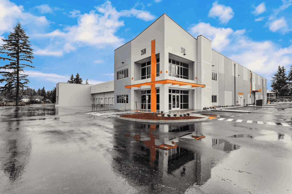 Screenshot 2026-02-26 131536 A modern two-story commercial building with gray and white walls and orange accents is shown under a bright blue sky. The wet pavement reflects the building and sky, and trees are in the background.