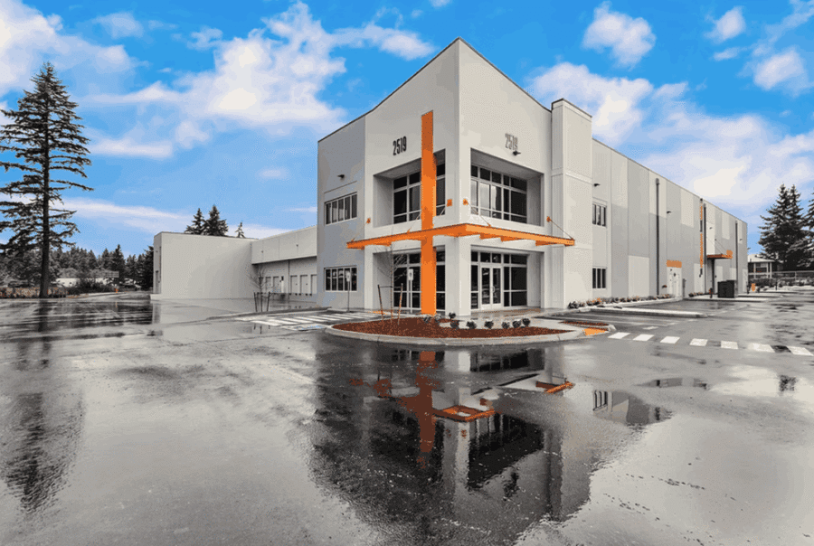 A modern two-story commercial building with gray and white walls and orange accents is shown under a bright blue sky. The wet pavement reflects the building and sky, and trees are in the background.