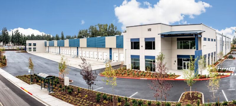 A large, modern warehouse designed by Seattle commercial architects features multiple loading docks, a blue and white exterior, landscaped surroundings, and a wide, empty parking lot under a partly cloudy sky.