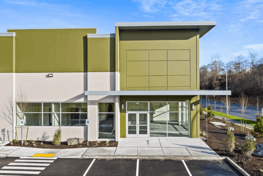 A modern commercial building by Seattle commercial architects, featuring olive and beige walls, large windows, and glass double doors, is surrounded by new landscaping and an empty parking lot under a blue sky.