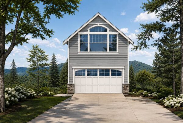 Gray two-story garage DADU with white trim and windows, featuring stone accents at the base, set among trees and mountains with a clear sky in the background. A concrete driveway leads up to the building, which showcases a practical DADU floor plan for versatile living or working space.