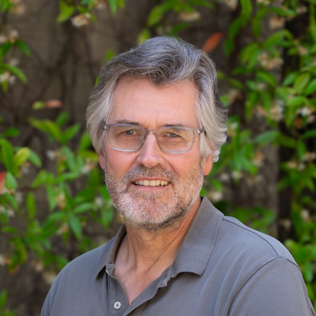A man with gray hair, glasses, and a beard is smiling while standing outdoors in front of green leafy bushes, wearing a gray collared shirt—his relaxed demeanor reflecting the creative spirit of an architecture firm professional.